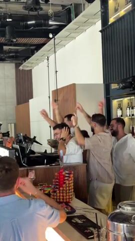 A group of men are dancing and cheering at a party. The DJ is at the decks with some equipment. Several people are holding cameras up to take pictures and videos. The environment appears to be a bar or club with lighting and shelves of bottles in the background.
