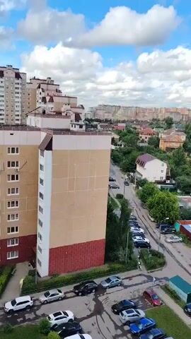 The video shows an aerial view of a residential area. In the foreground, there is a tall apartment building with a beige and red facade. Below the building, cars are parked in a lot and along a street. The street is lined with trees and smaller houses. In the background, there are more houses and apartment buildings stretching into the distance. The sky is blue with some white clouds. The overall scene depicts a typical urban neighborhood on a cloudy day.