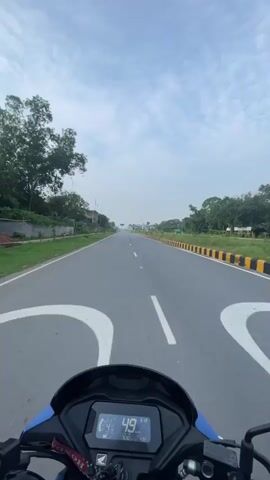 The video shows a first-person perspective of someone riding a motorcycle on a paved road. The rider's hands are visible on the handlebars, and the motorcycle's digital speedometer is displayed, showing speeds ranging from 49 to 58 kilometers per hour. The road is wide with a white dashed line marking the center and a yellow and black striped barrier on the right side. Trees and some buildings are visible along the sides of the road, and the sky is cloudy. The video appears to be recorded during daylight.