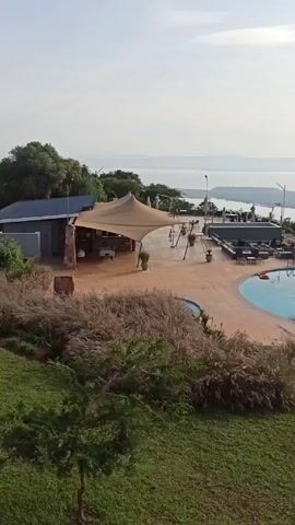 From an elevated perspective, a tranquil scene unfolds, showcasing a leisure area with a swimming pool and dining setup. The foreground is filled with lush greenery and tall grasses, leading to the immediate area around the pool which is adorned with lounge chairs and parasols. Underneath a large canopy, tables and chairs suggest a dining or relaxation space. In the distance, a vast expanse of water stretches to the horizon. The sun's rays reflect off the water's surface, creating a shimmering effect under a slightly cloudy sky. The overall atmosphere is peaceful and inviting, indicative of a resort or private retreat setting.