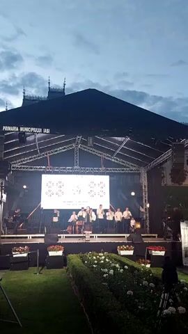 A group of musicians are performing on a stage outdoors. The stage is covered by a dark awning and illuminated by stage lights. A large screen behind the musicians displays a pattern. The musicians are dressed in traditional clothing and are playing instruments. In front of the stage, there is a manicured garden with flowers. In the background, there are buildings and a cloudy sky. The video is taken from a low angle, looking up at the stage.