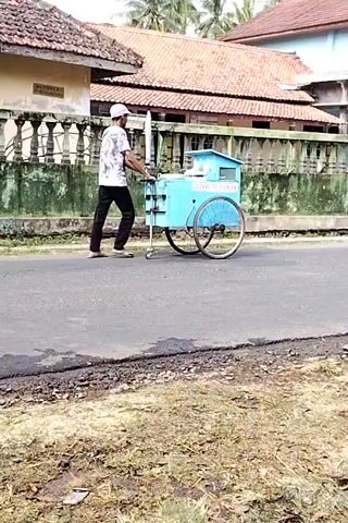 A person wearing a white cap, a white t-shirt with a black design, and black pants pushes a light blue cart with two large wheels down a paved road. The cart has a smaller wheel in front and a handle for pushing. The cart has text on its side that is difficult to read. The person is walking from left to right in the frame. In the background, there is a building with a red tile roof and a green fence. The ground next to the road is covered in dry grass and dirt. The overall scene appears to be outdoors during the daytime.