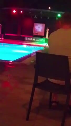The video shows a person sitting on a chair looking at a stage with a screen. The stage is lit with colorful lights, and there is a pool next to it. The person is wearing a white shirt and shorts. The video then pans to a blurry shot of people sitting at tables in an outdoor setting at night. One person is wearing orange pants and is looking at their phone. The setting appears to be a resort or hotel with outdoor seating and lighting.