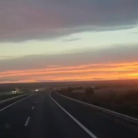 This is a first-person perspective video from inside a vehicle traveling on a highway during sunset. The sky is filled with dramatic colors, transitioning from deep purple and gray clouds at the top to vibrant oranges and yellows near the horizon. The sun is low, casting a warm glow over the landscape. The highway stretches ahead, with lanes marked by white lines. On either side of the road, there are fields and distant hills. The video captures the feeling of a journey through a beautiful, fading light.