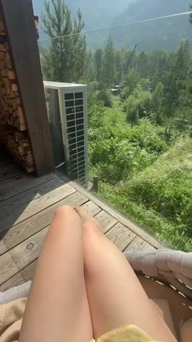 The video opens with a close-up of a person's legs resting on a wooden deck, with a view of lush green mountains and trees in the background. A white air conditioning unit is visible on the deck, as is a stack of firewood. The camera slowly pans up, revealing more of the mountainous landscape under a clear blue sky. The overall impression is one of a serene and relaxing outdoor setting.