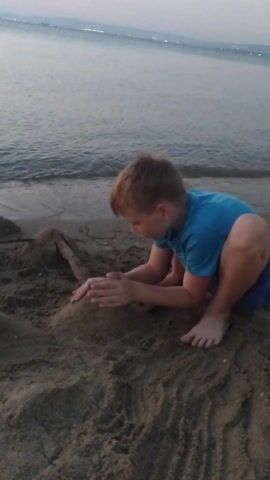 This media file captures a young child playing in the sand on a beach. The child, wearing a blue t-shirt and blue shorts, is crouched down, actively engaged with the sand, seemingly building something or playing with it. Their knees are drawn up to their chest, and their hands are outstretched, touching the sand.

The setting is a beach, with the calm sea visible in the background. The water appears a muted grey-blue, suggesting it might be overcast or nearing dusk. The sand itself has patterns and textures, indicating it has been disturbed by the child's play. A small stick or piece of driftwood is embedded in the sand near the child's hands, possibly incorporated into their sand creation.

The lighting suggests it is late afternoon or early evening, given the soft, diffused light. The overall mood appears peaceful and focused, with the child engrossed in their activity. The location is identified as Neoi Epivates, Greece. There is no visible text in the media file.