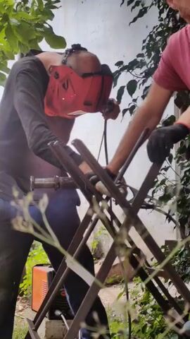 The video captures two individuals outdoors, engaged in a welding activity. The foreground is dominated by dark metal structures, possibly part of a project under construction, with some greenery obstructing the view. In the center, a person wearing a red welding mask and protective clothing is actively welding, sparks emanating from the point of contact. Their posture suggests focused effort. To the right, another person, wearing a pink shirt and black gloves, appears to be assisting or observing the welding process, with their hand resting on one of the metal structures. In the background, a white wall and lush green foliage are visible, indicating an outdoor setting. An orange welding machine is placed on the ground to the left, connected to the welding equipment. The lighting suggests it is daytime, with natural light filtering through the leaves.