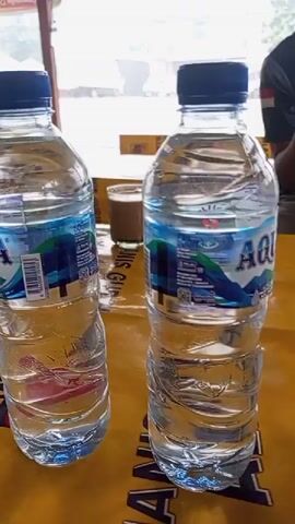 The video shows a close-up, slightly shaky, panning shot of several plastic water bottles. The bottles are all the same brand, "AQUA," with blue caps and clear water inside. They are arranged on a yellow surface, possibly a table, with some text or a pattern visible underneath. In the background, out of focus, there are glimpses of an outdoor setting, possibly a street or a market stall, with some blurry structures and what looks like a truck or vehicle. The lighting appears to be natural, suggesting an outdoor or well-lit indoor environment. There are no people clearly visible in the frame.