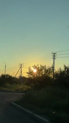 This video captures a view from a vehicle driving on a road. The sun is setting or rising, casting a warm glow and lens flare across the scene. Several utility poles with power lines are visible. There are various road signs, including a circular sign with the number "20" and a triangular sign indicating a speed bump. Lush green vegetation and trees line the side of the road. The sky is a clear, pale blue transitioning to a soft yellow near the horizon. The road surface appears to be asphalt, with some gravel and debris visible on the edge. The overall impression is one of a rural or semi-rural landscape bathed in the warm light of the sun.