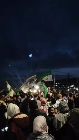 The video shows a large group of people, primarily seen from the back, gathered at an event under a dark, cloudy sky, possibly at night. Many are holding up Syrian flags, and some are recording the event on their cell phones. A person is being carried on someone's shoulders in the center of the crowd, waving a flag with Arabic script. The crowd is dense, and the atmosphere appears celebratory. There are bright lights shining from a stage or area in the distance.