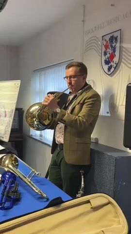 A man is playing a French horn in a room. He is wearing glasses, a brown checkered jacket, a patterned shirt, and green pants. He is standing next to a wall with a logo that says "ROCKWELL MUSIC ACADEMY". In front of him, on a blue cloth, are several brass instruments, including a trombone and a blue trumpet. A French horn case is visible in the foreground. There is a whiteboard with musical notes on it to the left of the man. The room appears to be a music classroom or practice space.