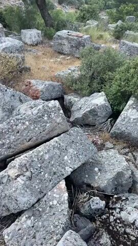 The video shows a rocky, uneven terrain with scattered large grey stones and some vegetation. The stones appear to be building materials, possibly remnants of an ancient structure. In the background, there are hills covered with trees and shrubs, and the sky is clear and blue. The lighting suggests it is daytime. There are no people or animals visible in the video. The camera movement is slow and panning, giving a sense of exploration or observation of the area. The overall impression is one of a somewhat wild, natural landscape with signs of past human presence.