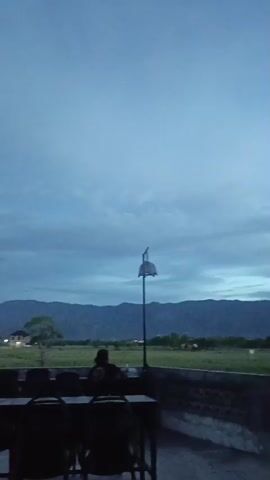 The video shows a dusk scene with a person sitting at a table outdoors, looking towards a landscape of fields and mountains. The sky is cloudy and a pale blue, indicating either early morning or late evening. A lamppost with an unusual umbrella-shaped top stands in the middle ground. In the foreground, several chairs and tables are visible, suggesting a seating area, possibly a cafe or restaurant patio. The overall atmosphere is calm and serene.