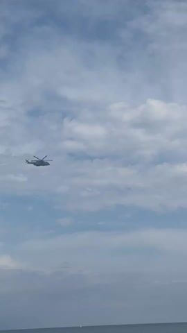 The video shows a grey helicopter flying in a cloudy sky above a body of water. The helicopter has multiple rotor blades and is seen from a distance. The clouds are white and grey, and the water appears calm.