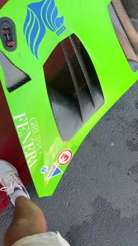 A bright green car hood, adorned with blue abstract designs and sponsor logos, is shown from multiple angles. It has vents made of carbon fiber and appears to be part of a race car. In some shots, the car's engine bay is visible, revealing complex mechanical components. The hood is placed in what seems to be a garage or pit area, indicated by the presence of tires, equipment, and a red floor. A person's leg and foot, wearing a white sneaker, are visible in the lower left corner of one frame. Another frame shows a wider view of the pit area with a large red garage door.