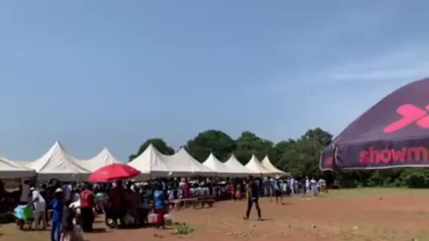 The scene appears to be an outdoor gathering, possibly a festival or event, with many people congregated under rows of white tents. A red umbrella is visible among the tents. A "Showmax" branded umbrella stands to the right of the frame. The ground is a mix of grass and bare earth. The sky is clear and blue, with some trees visible in the background. People are mostly standing and walking, suggesting movement within the event.