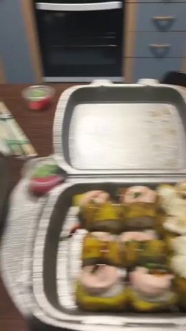 The video begins with a close-up of a dark wooden table. In the foreground, there is a white takeout container filled with food, possibly pasta with cheese. To the right, a partially opened plastic bag contains more food items. Further back on the table, there are plates, a glass of amber liquid (likely beer), and several other containers of food, including a small blue object that may be a phone or a small electronic device. The background shows kitchen cabinets and a countertop. The camera appears to be handheld and is moving, causing a blurred effect on the objects. There are no people clearly visible in the frame. The overall scene suggests a casual meal or gathering.