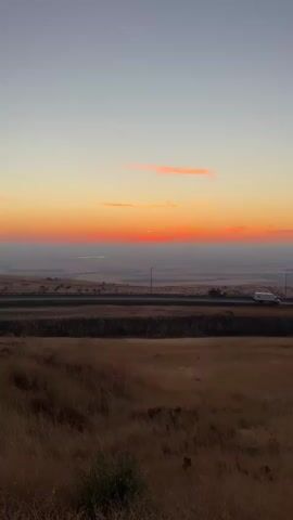 The video shows a wide-angle view of a landscape at sunset. The sky transitions from a light blue at the top to a vibrant orange and red near the horizon. There are a few wispy pink clouds scattered across the sky. In the mid-ground, a highway runs horizontally across the frame with a white vehicle visible on it. The foreground is a field of dry, golden grass. The overall impression is of a serene, natural scene.