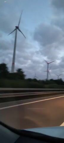 The video shows a view from a moving vehicle on a highway at dusk. Several wind turbines are visible on a hillside to the left of the road. The sky is overcast with dark clouds. The road has multiple lanes and a concrete barrier separates the lanes. The headlights of the vehicle are on, casting reflections on the wet road surface. The wind turbines are tall, white structures with three blades each. Some of the turbines have red lights blinking at the top. The overall mood of the video is somber and atmospheric due to the low light conditions and the cloudy sky.