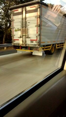 A truck is visible on a road through a window. The truck is white with red and yellow markings. It appears to be a delivery truck with a large cargo container. The truck is in motion, and the camera appears to be in another vehicle traveling alongside it or behind it. The road is paved and appears to be a highway or a similar main road. Trees are visible in the background, suggesting an outdoor environment. The lighting indicates it is daytime, possibly late afternoon due to the angle of the sun.