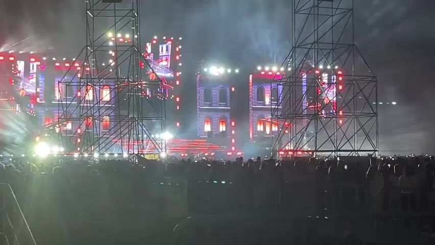 The video captures a nighttime outdoor concert with a large crowd gathered in front of a stage. The stage features a elaborate set design resembling a castle or large building, illuminated with red and purple lights and smoke effects. Two large metal scaffolding structures flank the stage. The audience, a dense mass of people, is visible in the foreground, with some security personnel also present. The atmosphere appears energetic with lights and pyrotechnics contributing to the spectacle.