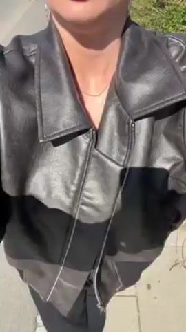 The video shows a person, from the neck down, wearing a black leather jacket and black pants. The jacket is zipped halfway up. The person is wearing a gold necklace. The setting appears to be outdoors, with what seems to be a sidewalk and green vegetation visible in the background. The camera angle suggests the video is being recorded from the person's perspective, looking down.