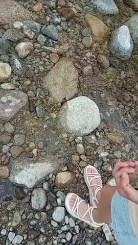 This is a first-person point-of-view video of a person's feet wearing white sandals standing on a rocky riverbed. The water is shallow and clear, flowing over the rocks. The person is holding a small rock in their hands and appears to be examining it. The scene is natural and outdoors.