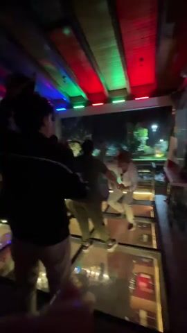 The video captures a lively scene at a venue with a unique glass floor, possibly a restaurant or club, at night. A group of people are enthusiastically dancing, bathed in the colorful glow of red, green, and blue lights illuminating the wooden ceiling. In the foreground, three individuals are visible, with the person on the far left turned away from the camera, wearing a dark jacket and light-colored pants. The central figure, also facing away, is wearing a dark olive shirt and light pants. The individual on the right is wearing a white outfit and a cowboy hat, actively dancing with their arms outstretched.

As the video progresses, more people join the dance floor. The glass floor reveals a lower level, where bottles and glasses are visible, suggesting a bar or storage area beneath. The background shows a glimpse of the outdoors through a glass wall, with trees and a lamp post visible, indicating it is nighttime. A Mexican flag is prominently displayed on the right side of the frame. The atmosphere appears festive and energetic, with the dancers exhibiting joy and excitement. The location cues, like the Mexican flag and the general ambiance, align with the provided context of Ocoyoacac, Mexico. There is no visible text within the video.