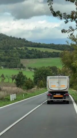 The video shows a rear view of a white dump truck driving on a paved road. The truck is on the right side of the road, moving away from the camera. The road has white dashed lines on the right side and solid white lines on the left side. The truck is carrying a load of dark material in its bed. The surrounding environment is a rural landscape with rolling green hills, trees, and fields. The sky is cloudy with some patches of blue. The overall atmosphere is serene and suggests a travel or scenic journey.