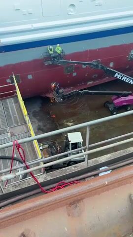 Two workers stand on a boom lift painting the side of a ship that is mostly red, with the top part painted gray and a blue strip above the gray. A third worker stands in a boom lift with sparks flying from the side of the ship. The ship sits in a dry dock area with a flatbed truck visible and a yellow scaffold with red cords.