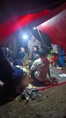 Inside a red-tented shelter, a group of people are gathered. At least five individuals are visible, sitting on a patterned rug spread over the ground. The tent is illuminated by a bright, hanging light, with a second light casting a red hue from above. The people appear to be engaged in conversation, some smiling. Around them, there are bags, cups, a kettle, and other personal items scattered on the ground. The atmosphere is intimate and casual. The ground outside the rug is uneven and looks like dirt or grass.