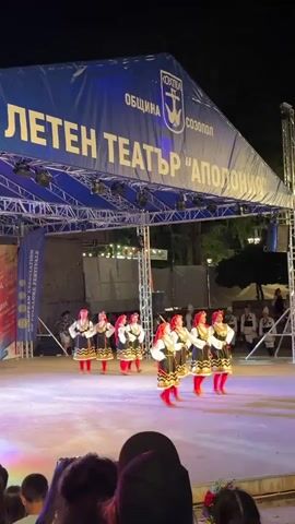 The video shows an outdoor performance on a stage at night.  A large blue banner with white text is visible above the stage. The text on the banner includes "ЛЕТЕН ТЕАТЪР 'АПОЛОНИЯ'".  Below the banner, there is a stage with theatrical lighting.  On the stage, a group of people, appearing to be female dancers, are performing. They are wearing traditional folk costumes, which include dark vests over white blouses, long skirts with red accents, and red leg coverings. They are arranged in a line and are performing synchronized movements.  To the left of the stage, a vertical banner with text and an image is displayed.  In the background, there are trees and some lighting structures.  In the foreground, the backs of audience members are visible, indicating a live audience watching the performance.  The stage floor is illuminated with colored lights, primarily white and pinkish hues.