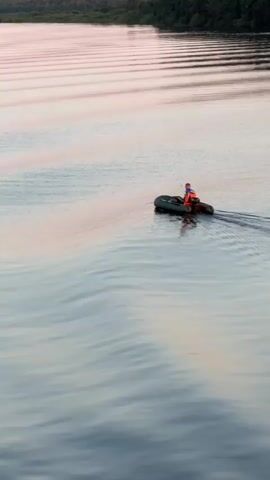 A person wearing an orange life vest sits in an inflatable boat moving across a calm body of water. The boat is leaving a wake. The water reflects the soft colors of the sky, with gentle ripples creating patterns on the surface. In the distance, a treeline borders the horizon under a pinkish-purple sky.
