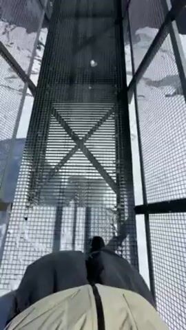 The video shows a first-person perspective of someone walking on a metal grate walkway, surrounded by a metal cage. The walkway leads to a platform with a sign that reads "La Clusaz". The surrounding environment is a snow-covered mountain range under a clear blue sky. The sun is visible in the sky.