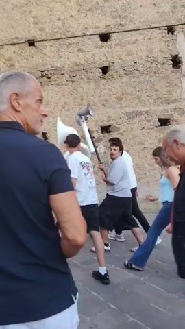 The video shows a street scene with several people. In the foreground, a man with graying hair wearing a dark blue polo shirt is visible from behind. To his right, another man with white hair wears a red vest over a blue shirt and jeans. In the background, a group of people are walking. One man wears a light grey shirt and jeans, while another wears a white shirt and black pants. Further in the background, people are seated on a bench and others are walking in a plaza. The buildings in the background appear to be old stone structures with balconies. A small tree is planted in a large planter on the left side of the plaza. The ground is paved with large stone tiles.