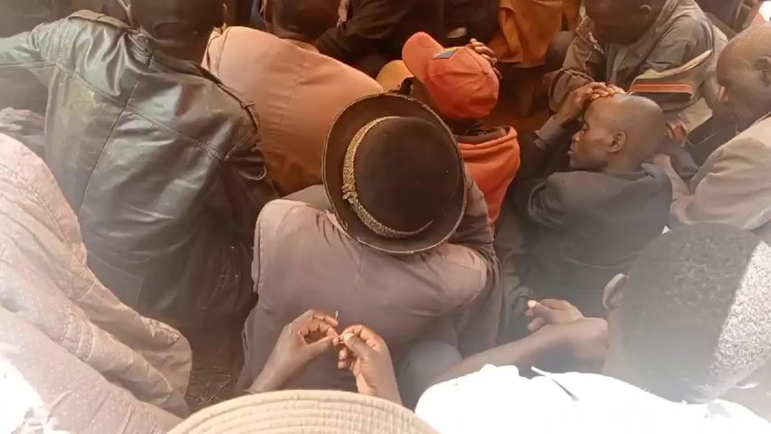 A large group of men are gathered together outdoors, sitting on chairs and the ground under a tarpaulin shelter. Many are wearing hats, including a black fedora, a red baseball cap, and several other styles. The men are mostly dressed in casual or semi-formal attire, including jackets, shirts, and pants. Some individuals are standing on the periphery of the group. There are two open umbrellas, one white and one pink, held by individuals standing further back. The ground appears to be soil with some greenery. The overall impression is a gathering or meeting of some kind.