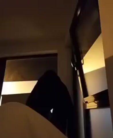 The video depicts a dimly lit room with a partially visible bed in the foreground. A person, shrouded in dark clothing, stands near a window. The window is partially covered by blinds, with light filtering through. A television is mounted on the wall near the window. The person remains largely still throughout the video.