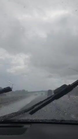 The video is a first-person perspective from inside a vehicle driving on a highway during a rainstorm. The windshield is covered in raindrops, distorting the view. The highway is wet, and several cars are visible ahead, driving in the same direction. The sky is overcast with dark, heavy clouds. The overall atmosphere is gloomy and wet due to the rain.