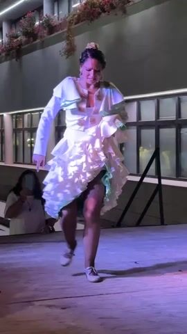 A woman in a ruffled white flamenco dress and white sneakers dances on a wooden stage. She is moving her arms and her dress is swaying. Her hair is pulled up. A woman in a white shirt is standing in the background to the left, holding a phone. The background shows a building with windows and plants. The stage is lit with purple and pink lights.