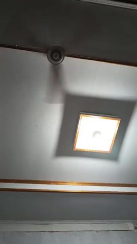 The video shows a ceiling with a square light fixture and a ceiling fan. The light is on, casting a shadow from the fan onto the ceiling. The fan is spinning, indicated by the blurred blades and the moving shadow. There are decorative wooden trim pieces along the ceiling.