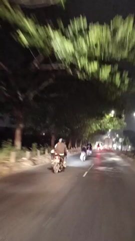 The video captures a night scene on a road in Wangunharja, Indonesia. The subjects are several people riding motorcycles. The road appears to be wet, suggesting it might have rained recently or is still raining lightly, and streetlights illuminate the path. Lush green trees line the left side of the road, with their leaves illuminated by the streetlights, creating a dappled effect. The overall mood is calm and perhaps a bit melancholic due to the nighttime setting and the quiet movement of the motorcycles. There is no visible text in the video.