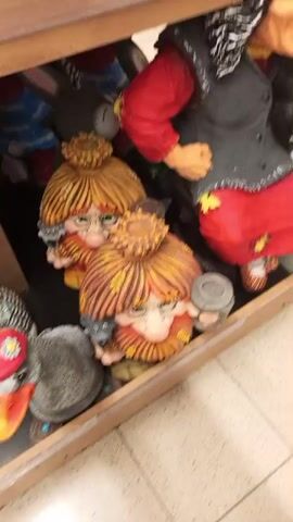 The video shows a shelf in a store filled with garden decorations. On the top shelf is a red squirrel figure and a garden gnome figure of an old woman with a pointy nose, red skirt, black vest, and a blue hat. The figure also appears to have a cat. On the shelf below, are a pair of gnome figures with orange straw-like hair and glasses. The next shelf down has garden decorations of gray rabbits and an owl.