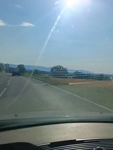 The video shows a view from inside a vehicle traveling on a road on a sunny day. The camera is pointed forward, capturing the road ahead and the surrounding landscape. The sun is shining brightly, creating lens flares across the windshield. The road is paved with white lines marking the lanes. In the distance, a truck is visible on the road. The landscape on either side of the road includes green fields, trees, and hills. There are also some tiered structures that appear to be part of an agricultural setup or some kind of infrastructure in the fields. The sky is blue with a few white clouds. The interior of the car is visible at the bottom of the frame, including the dashboard and steering wheel. The video appears to be a short clip of a journey.