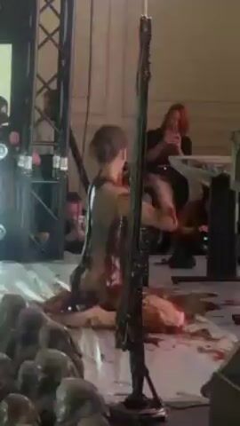 A woman is lying on the floor, apparently covered in fake blood, with people surrounding her and taking photos. There are stage props and equipment visible in the scene. The lighting is primarily red.