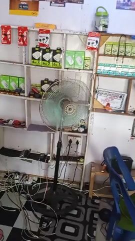 A pedestal fan is positioned in front of shelves filled with electronic accessories, including phone chargers and earbuds. The fan is in motion, circulating air in a cluttered room. Numerous cables are strewn across the floor and plugged into power strips, suggesting a charging station or repair area. Two chairs, one blue and one wooden, are visible in the background. The shelves display various products, some in their original packaging, indicating a retail or service environment. The overall scene suggests a busy, functional space dedicated to electronic devices.
