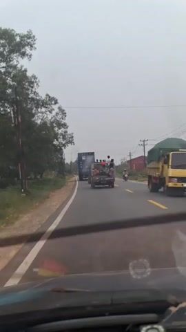 The video shows a view from inside a vehicle, looking out through the windshield. The vehicle is on a paved road with a white line on the left side and a yellow line down the center. Trees and vegetation line the left side of the road, and some buildings and power lines are visible on the right. Several vehicles are on the road. In front, there is a truck with people in the back. Further ahead, there is a larger truck. A motorcycle is also visible on the right side of the road. The sky is overcast. The video captures movement as the vehicle drives along the road.