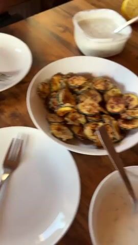 The video pans across a wooden table laden with food, suggesting a meal setting. In the foreground, a white bowl contains fried zucchini slices. To the right, a white takeout container is divided into two compartments, one filled with what appears to be stewed fruit and the other with bread cubes. A smaller white bowl with a spoon rests in front of this container, filled with a creamy dip, possibly ranch dressing. In the background, out of focus, another dish with a dip and a lemon wedge are visible. Further back, on a wooden surface, are two small decorative vases with flowers. The camera then tilts up to reveal a television screen displaying an image of two women, possibly from a music video or photoshoot. The room appears to be furnished with wooden walls and a bed with white linens.