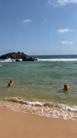 The video shows a beach scene with two children wearing yellow life jackets wading in shallow water. The water is clear with small waves. A rock formation is visible in the water further out. The beach has light brown sand. The sky is blue with some scattered white clouds. The scene is shot from the beach, showing the shoreline and the children enjoying the water. One child briefly floats on their stomach.
