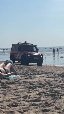 A red and white vehicle, likely a rescue or emergency response vehicle, is parked on a sandy beach near the water's edge under a clear sky. Several people are visible in the background, some wading in the shallow water and others standing on the beach. In the foreground, a person's legs are visible, reclining on the sand. A young person in a leopard-print bikini walks across the sand in front of the vehicle. The vehicle has lights on its roof and a Dutch license plate. The overall scene depicts a sunny day at the beach with a sense of normalcy, interrupted only by the presence of the rescue vehicle.