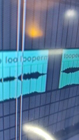 The media file displays a close-up view of a digital audio workstation (DAW) interface, likely on a computer screen. The screen shows two audio tracks, each represented by a turquoise-colored waveform visualization within a grid-like timeline. The waveforms appear to be audio clips, possibly loops, as suggested by the text visible on them. The text, oriented vertically, reads "loopern".

The scene is dimly lit, with a soft, yellowish light emanating from the left side of the frame, casting a blurred glow on the screen. There are also bright, out-of-focus reflections on the screen, including a white circular glare on the upper left and a curved white glare on the right. These reflections suggest ambient lighting conditions or potentially light sources near the screen. The grid pattern of the DAW interface implies a structured digital environment, likely a home studio or a production space.

No people are visible in the frame. The activity taking place is the display of audio editing software, with two audio clips loaded and ready for manipulation. The overall mood is neutral and technical, focused on the visual representation of sound data. The location context of Remedios de Escalada, Argentina, does not provide specific visual cues within the image itself, as the image is a close-up of a digital interface. The time of day and weather are not discernible from the provided image.